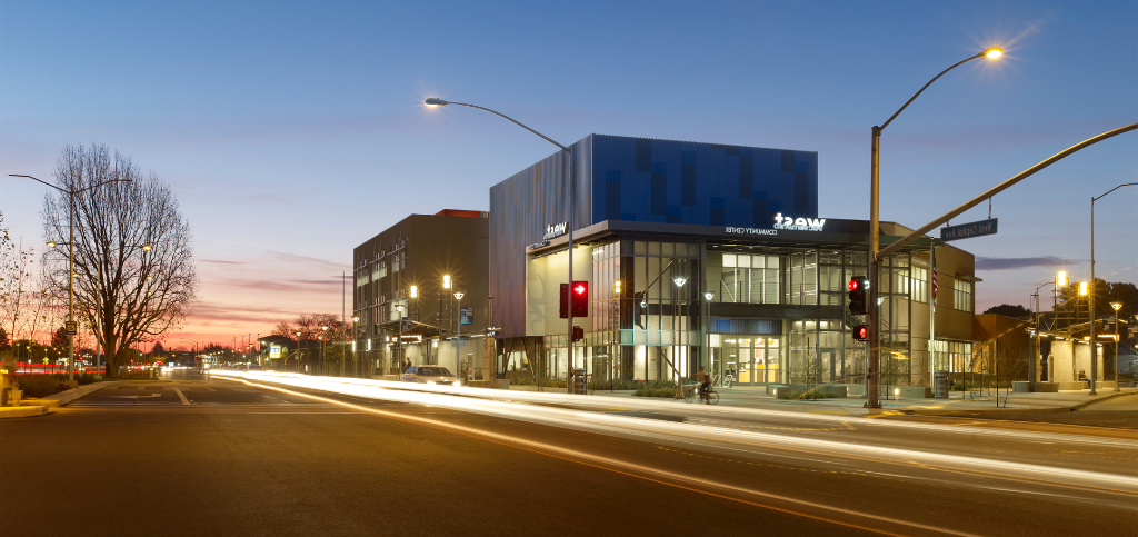 A wide shot of the Community Center building during the evening, a flash of blurred headlights transcends through the street in front of the building.